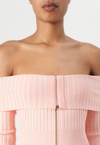 Ribbete off-shoulder topp i myk rosa, med en gullglidelås foran. Tøyet har en glatt tekstur med vertikale linjer.