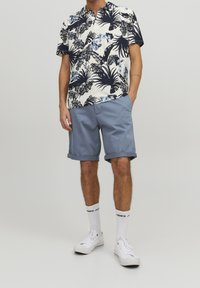 Camicia a maniche corte con colletto in bianco, decorata con stampa floreale blu e palme, abbinata a pantaloni corti azzurri e sneakers bianche con dettagli neri.
