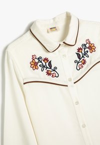 Blusa cremosa con cuello, que presenta ribetes en marrón oscuro y colorido bordado floral en el pecho. Tejido ligero y suave; cierre de botones.