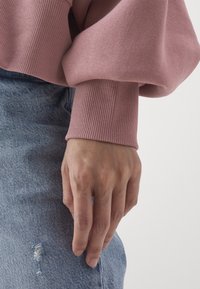 Main détendue le long du corps, portant un pull rose à manches longues avec poignets côtelés et un jean bleu déchiré, sur un fond uni.