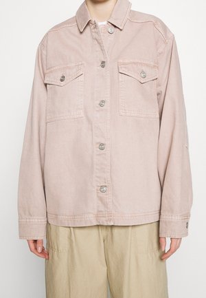 Persoon die een lichtroze katoenen jas met knopen draagt, met twee klepzakken aan de voorkant en beige broek, gefotografeerd van nek tot heupen tegen een witte achtergrond.