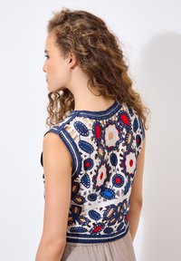 Häkel-Crop-Top mit floralen Mustern in Rot, Blau und Beige, ausgestattet mit einem marineblauen Saum. Das Rückendesign zeigt offene Maschen und dekorative Details.