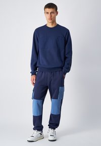 Felpa blu navy con dettaglio sul pannello frontale, abbinata a pantaloni jogger blu navy con toppe azzurro chiaro e tasche laterali. Sneakers bianche completano il look.