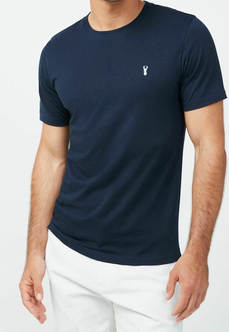 Homme portant un t-shirt bleu marine à manches courtes avec un petit logo de cerf blanc sur la poitrine et un pantalon blanc.
