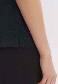 Tessuto della t-shirt verde scuro con una trama sottile, dotata di orlo arrotondato e aperture laterali. Si abbina a pantaloni neri.