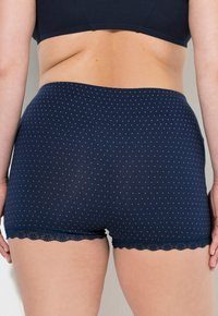 Lato inferiore della schiena e fianchi di una donna che indossa mutande tipo boyshort a pois blu navy con bordi in pizzo, su uno sfondo chiaro e semplice.