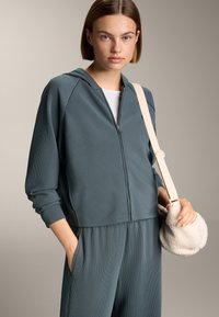 Felpa teal a coste con zip e cappuccio, abbinata a pantaloni coordinati. Il modello porta una piccola borsa texturizzata color crema.