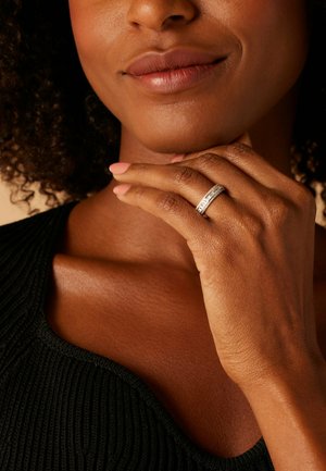 Silver ring with small, clear stones set in a simple band, worn on a hand with tanned skin against a black textured top.