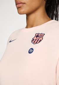 Svijetlo roza pamučna majica s izvezenim logotipom FC Barcelone u tamnoplavoj i roza boji, s plavim logotipom Spotify otisnutim ispod. Okrugli izrez.