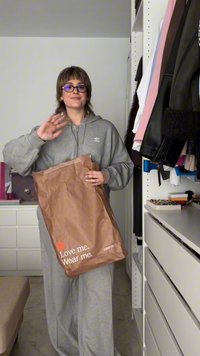 Grå oversized huva med ficka fram, hålls av en person. Brun papperspåse med vit text "Älska mig. Bär mig." synlig.