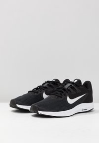 Chaussures de sport noires avec un dessus en maille texturée, un logo Nike blanc, des lacets noirs et une semelle blanche rembourrée. Design léger et flexible.