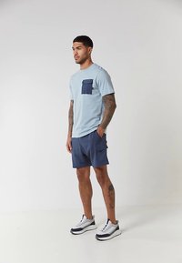 Muž s tetovaniami, oblečený v svetlomodrom tričku s tmavomodrým vreckom, námorníckych šortkách a bielych teniskách, stojí na jednoliatom pozadí.