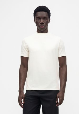 Hombre con el pelo corto y rizado, lleva una camiseta blanca lisa de manga corta y pantalones negros, de pie frente a un fondo liso y claro.