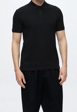 Homme portant un polo noir ajusté à manches courtes, avec un col et une patte de boutonnage à deux boutons, associé à un pantalon noir ample.