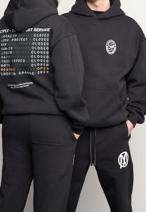 Dos personas que llevan sudaderas con capucha y pantalones negros; una sudadera muestra texto blanco y amarillo en la espalda, la otra tiene un logo circular en el pecho.