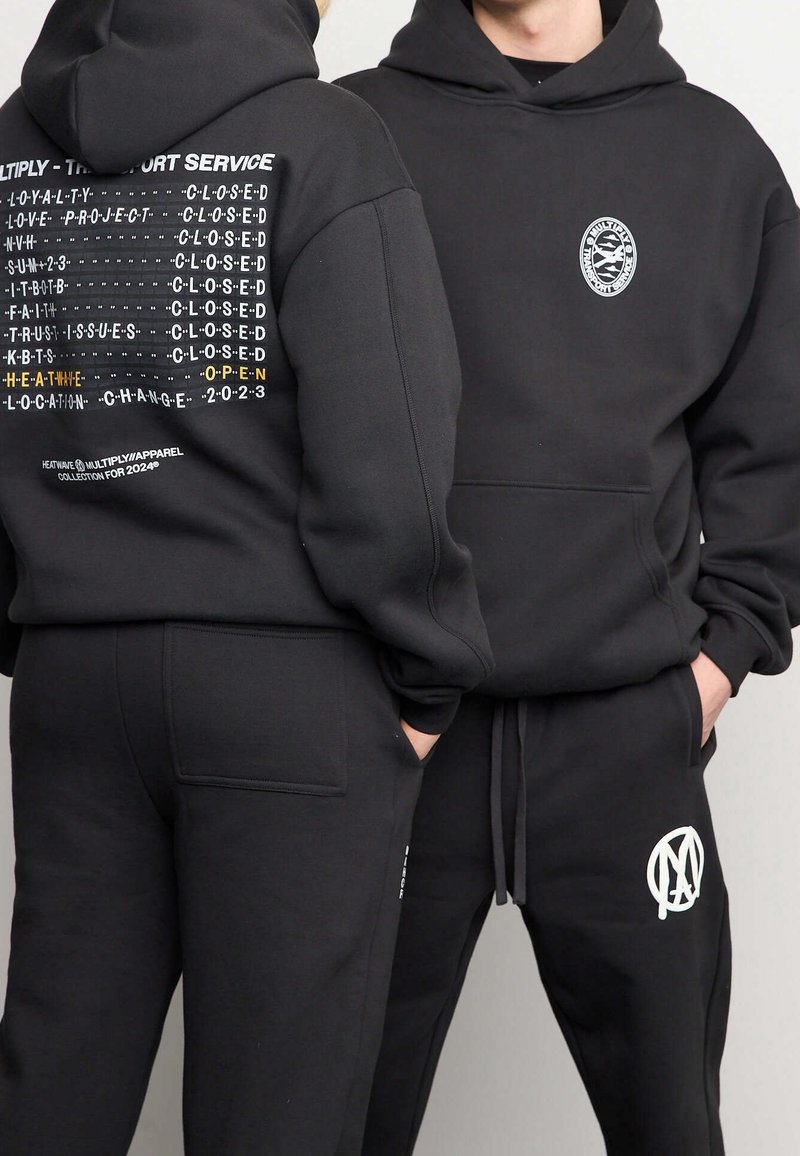 Deux personnes portant des sweats à capuche noirs et des pantalons ; un sweat à capuche affiche un texte blanc et jaune dans le dos, l'autre a un logo rond sur la poitrine.