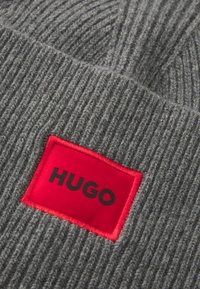 Berretto in maglia a coste grigio con una grande toppa rossa quadrata con il testo nero "HUGO". Tessuto ad alta consistenza con fibre visibili.