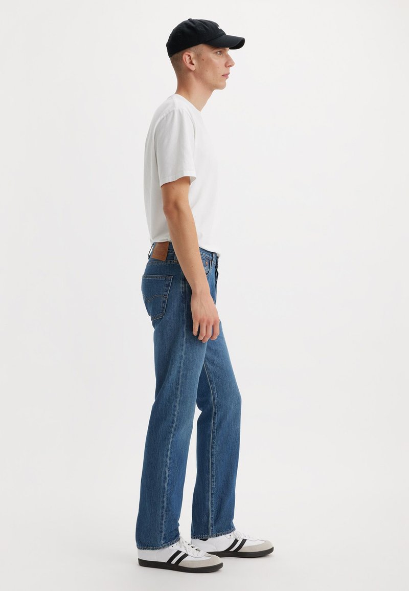 Blaue Jeans mit geradem Bein, kombiniert mit einem weißen T-Shirt und einer schwarzen Mütze. Die Schuhe sind weiß mit schwarzen Streifen. Einfacher, lässiger Stil.