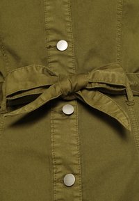 Camicia in tessuto verde oliva con cintura da annodare, bottoni a pressione argento e dettagli di cucitura in vista. Materiale in cotone texturizzato con una finitura liscia.