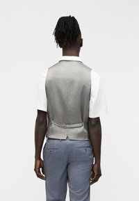 Άνδρας με ντραντλοκς, φοράει γκρι γιλέκο πάνω από λευκό κοντομάνικο πουκάμισο και μπλε παντελόνι φόρμας, φαίνεται από πίσω.