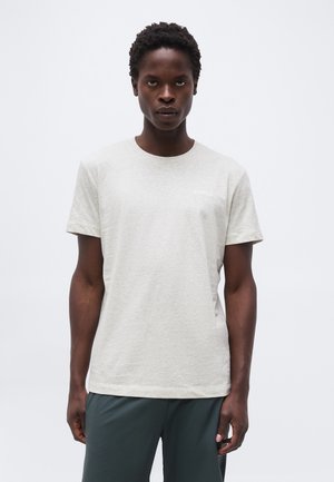 Νέος άνδρας με σκούρα σγουρά μαλλιά, που φοράει ανοιχτό γκρι κοντομάνικη μπλούζα και σκούρο πράσινο παντελόνι, μπροστά σε απλό λευκό φόντο.