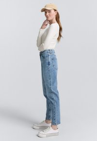Maglietta chiara a maniche lunghe, jeans azzurri a vita alta e sneaker bianche con strisce nere. Un cappellino color sabbia completa l'outfit.