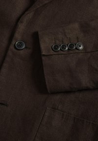 Manche et panneau avant d'une veste de costume marron foncé montrant les détails des boutons, y compris quatre boutons de manchette et un bouton à l'avant.