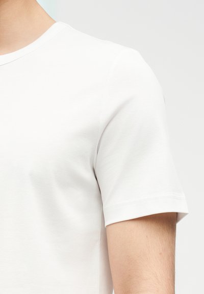 Κοντινό πλάνο ενός ατόμου που φοράει ένα απλό λευκό κοντομάνικο μπλουζάκι με στρογγυλή λαιμόκοψη, εμφανίζοντας λεπτομέρειες στον ώμο και το πάνω μέρος του μπράτσου.