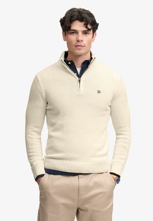 Ung mand iført en cremefarvet halv-lynlåstrøje over en navy skjorte, beige bukser og et armbåndsur, stående med hænderne i lommerne.