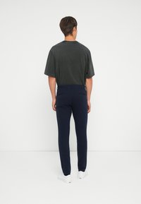 T-shirt in cotone grigio scuro con maniche corte, abbinato a pantaloni blu navy su misura. Sneakers bianche visibili ai piedi. Sfondo semplice.