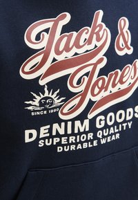 Logo de Jack & Jones en tejido navy con un gráfico del sol y el texto "Denim Goods Superior Quality Durable Wear Since 1990"