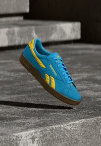 Modro-rumena Reebok superga, obešena nad sivimi betonskimi stopnicami, prikazuje stran in podrobnosti podplata.