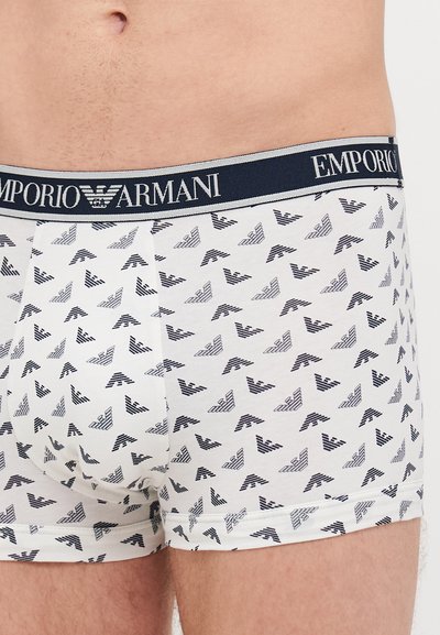 Bijele pamučne bokserice s ponavljajući uzorak loga orla u tamnoplavoj boji, s tamnoplavim pojasom na kojem je ispisano "EMPORIO ARMANI" u srebrnoj boji.