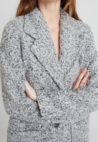 Harmaa teksturoitu kaksirivinen takki pehmeällä kankaalla, leveillä kauluksilla ja suurilla napeilla. Etutaskut ja rento istuvuus.