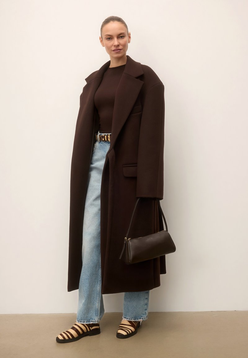 Femme portant un long manteau marron foncé, un haut marron, un jean bleu clair, des chaussures rayées, tenant un petit sac à main marron foncé contre un mur blanc.