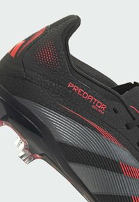Black soccer cleat with textured upper, red accents, and silver stripes. Features a high collar and transparent outsole with studs.