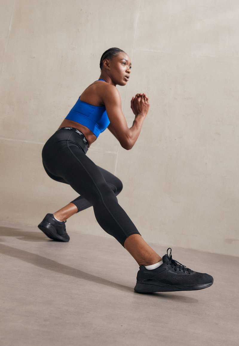 Zapatillas deportivas negras con malla texturizada, combinadas con leggins negros ajustados y un sujetador deportivo azul. El sujeto está en una posición de zancada dinámica.