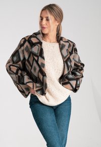 Braun und cremefarben gemusterte Jacke mit geometrischen Designs, taillierter Form und breiten Revers, getragen über einem cremefarbenen Strickpullover und blauen Jeans.