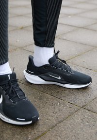 Ténis desportivos pretos com um tecido em malha texturizado, sola branca e logótipo da Nike. Combinados com leggings pretas riscada e meias brancas sobre uma superfície pavimentada.