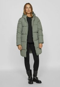 Manteau long matelassé en vert olive, avec une capuche, fermeture éclair à l'avant et poches latérales, associé à un haut et un pantalon noirs, et des bottes à boucle noires.