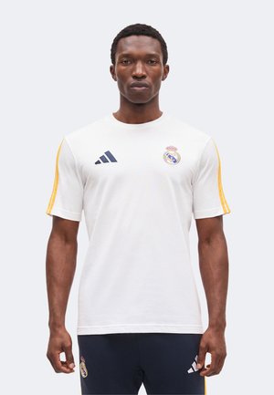 Bărbat purtând un tricou de antrenament alb al echipei Real Madrid cu dungi galbene pe mâneci și pantaloni bleumarin, stând pe un fundal simplu, luminos.
