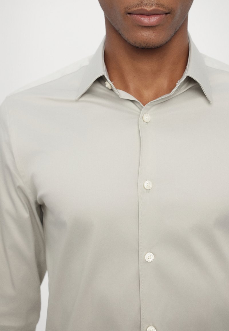 Chemise boutonnée gris clair avec une texture lisse, col pointu et boutons blancs. Design simple, coupe ajustée sans motifs ni accents.