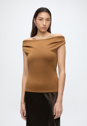 Femme aux longs cheveux foncés portant un haut marron décolleté aux épaules et une jupe noire sur un fond clair uni.