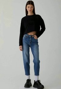 Sweatshirt preto cropped com texto em relevo, combinado com jeans azul claro, ligeiramente soltos. O modelo usa ténis pretos e meias brancas.