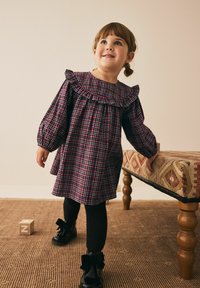 Karierenkleid in Lila, Schwarz und Weiß, mit einem Rüschenkragen und langen Ärmeln, kombiniert mit schwarzen Strumpfhosen und lackierten Schuhen mit Schleifen.