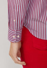 Camisa a rayas rojas y azules con un puño que cuenta con dos botones, combinada con pantalones de pana rojos visibles por la parte inferior. Textura suave.