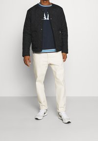 Giacca nera trapuntata con colletto rotondo, indossata sopra una camicia a righe blu navy, abbinata a pantaloni chiari e scarpe da ginnastica bianche.