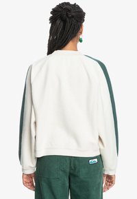Sudadera de forro polar en color crema con acentos en verde oscuro en los hombros, que presenta un ajuste relajado y un cuello de canalé. Combinada con pantalones de pana verdes.