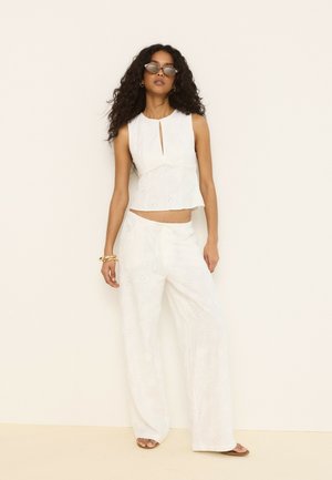 Femme aux longs cheveux bouclés portant un haut blanc sans manches et un pantalon large avec des détails à œillets, des lunettes de soleil et des bracelets en or, debout calmement.