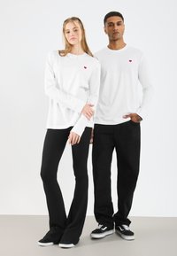 Λευκά πουκάμισα με μακριά μανίκια και μικρό κόκκινο λογότυπο καρδιάς, συνδυασμένα με μαύρα φαρδιά παντελόνια. Ένα μοντέλο φοράει αθλητικά παπούτσια, ενώ το άλλο έχει τα χέρια στις τσέπες.